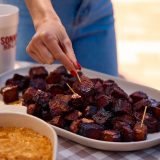 Woman grabs burnt ends by toothpick from platter